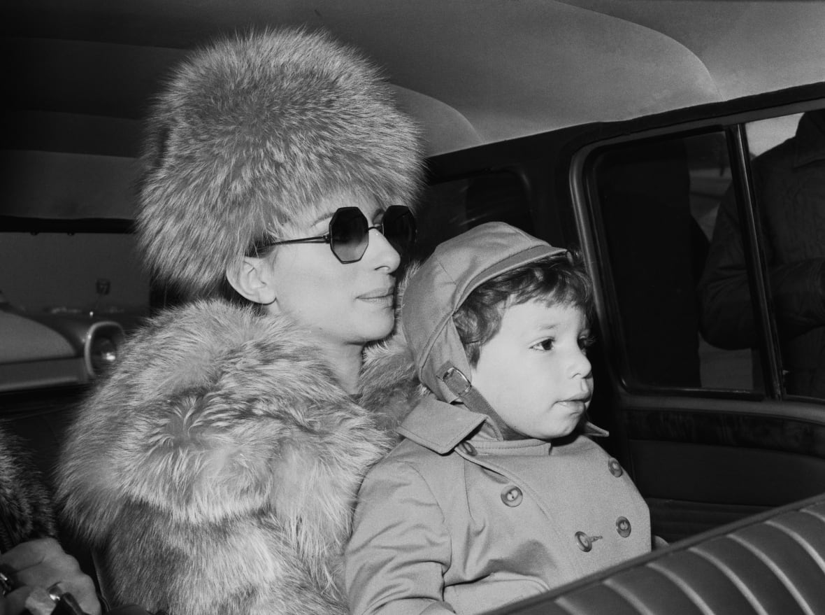 Black and white photo. Streisand, wearing a fur hat and coat, sits in a car with her son on her lap.