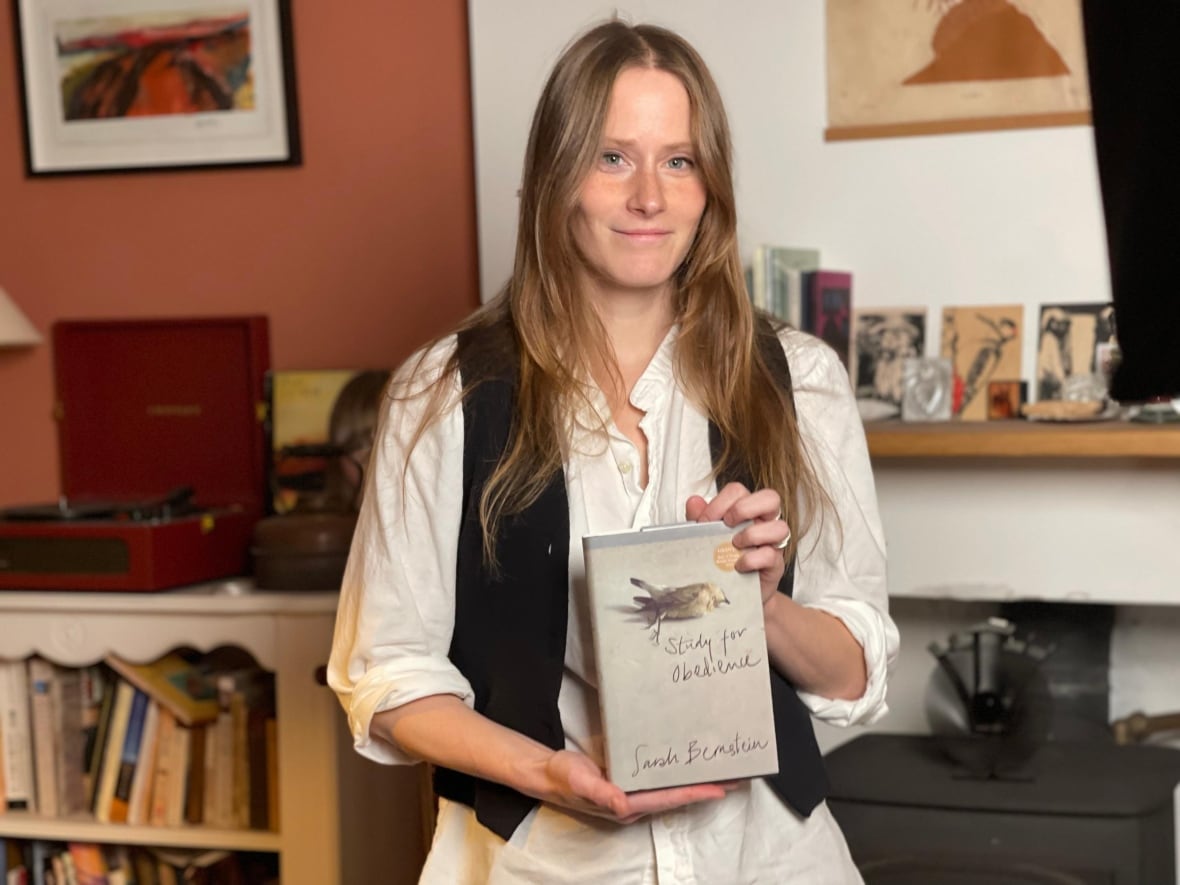 A woman with long auburn hair holds a book cover in her hands. She is standing in a living room with books in the background. 