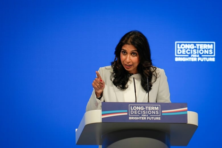 Une femme en blazer montre du doigt tout en s'exprimant sur un podium lors d'un événement public.