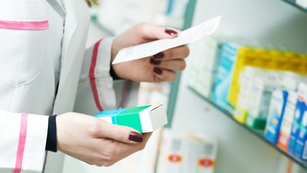 A supply chain delay and increased demand have led to a shortage of certain prescription painkillers in Canada. A closeup shows the hands of a woman in a white lab coat in a store, holding a prescription in one hand and a box of medicine in her other.