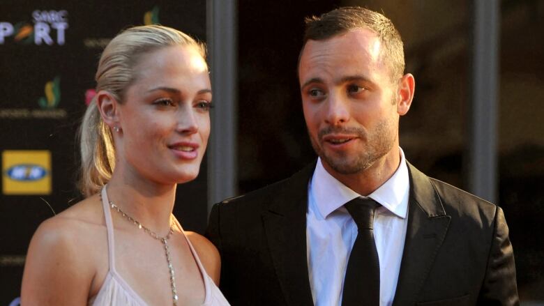 A man in a dark suit and tie and a woman in a sleeveless dress and necklace are shown together at a public event.