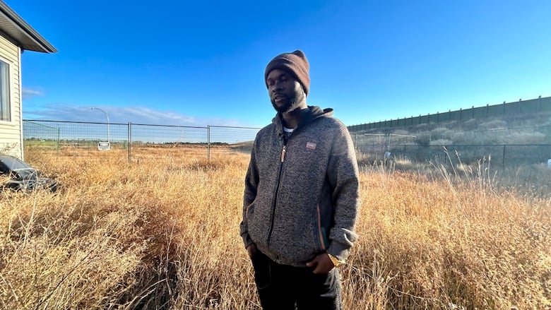A man in a grey sweater and black pants stands in an overgrown yard.