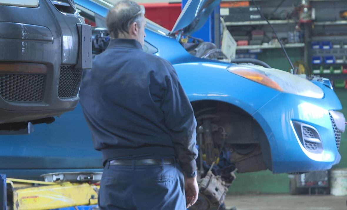 A person in a dark blue jumpsuit stands facing blue care hoisted up with a wheel removed.