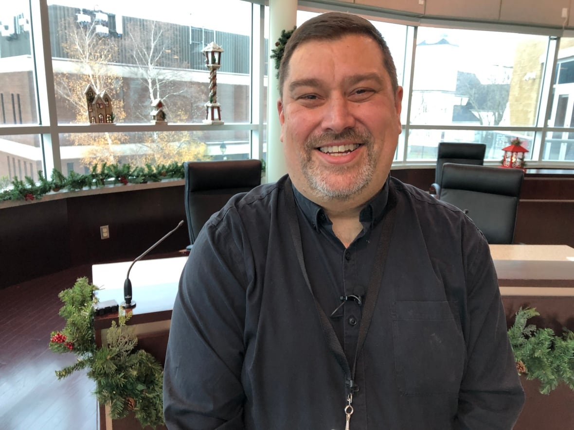 A smilling man wearing a black dress shirt stands in a city hall chamber. The chamber is decorated for Christmas.