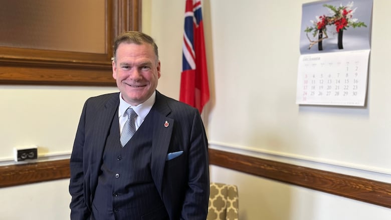 A man in a suit stands in front of a flag.