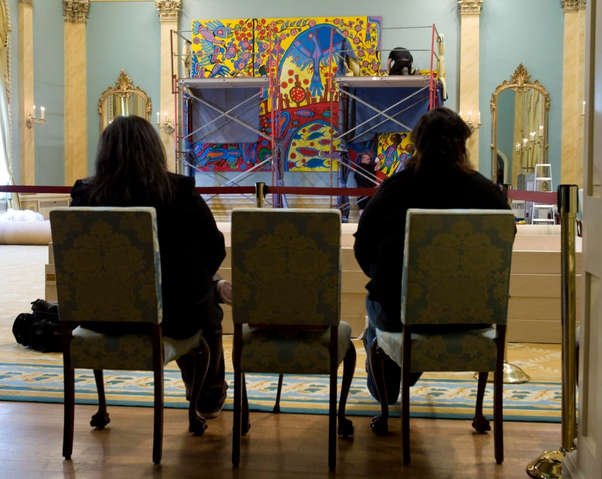 Two women sit in chairs in an ornate room. In front of them, workers on scaffolding are hanging a massive canvas on the wall.