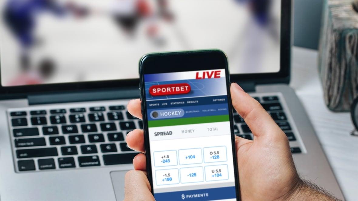 Someone holds a phone with an open hockey bet website in front of a laptop playing a hockey game.