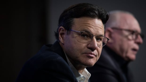 A white man with black hair and wearing glasses and a black suit stares into the camera. An older white man speaks behind him.