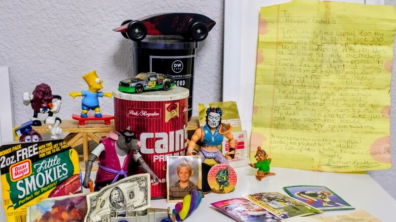 A hand-written letter on yellow paper, neatly propped up against the wall next to an old coffee tin, an Oscar Meyer Little Smokies label, a (fake) $50 bill, two toy cars, a wallet-sized photo of a little boy, a sticker, a piece of paper with printed words, a Pog and Slammer, some Play-Doh, two Ninja Turtle action figures, a figurine of a bear on a skateboard, two California Raisins and a Bart Simpson toy. 