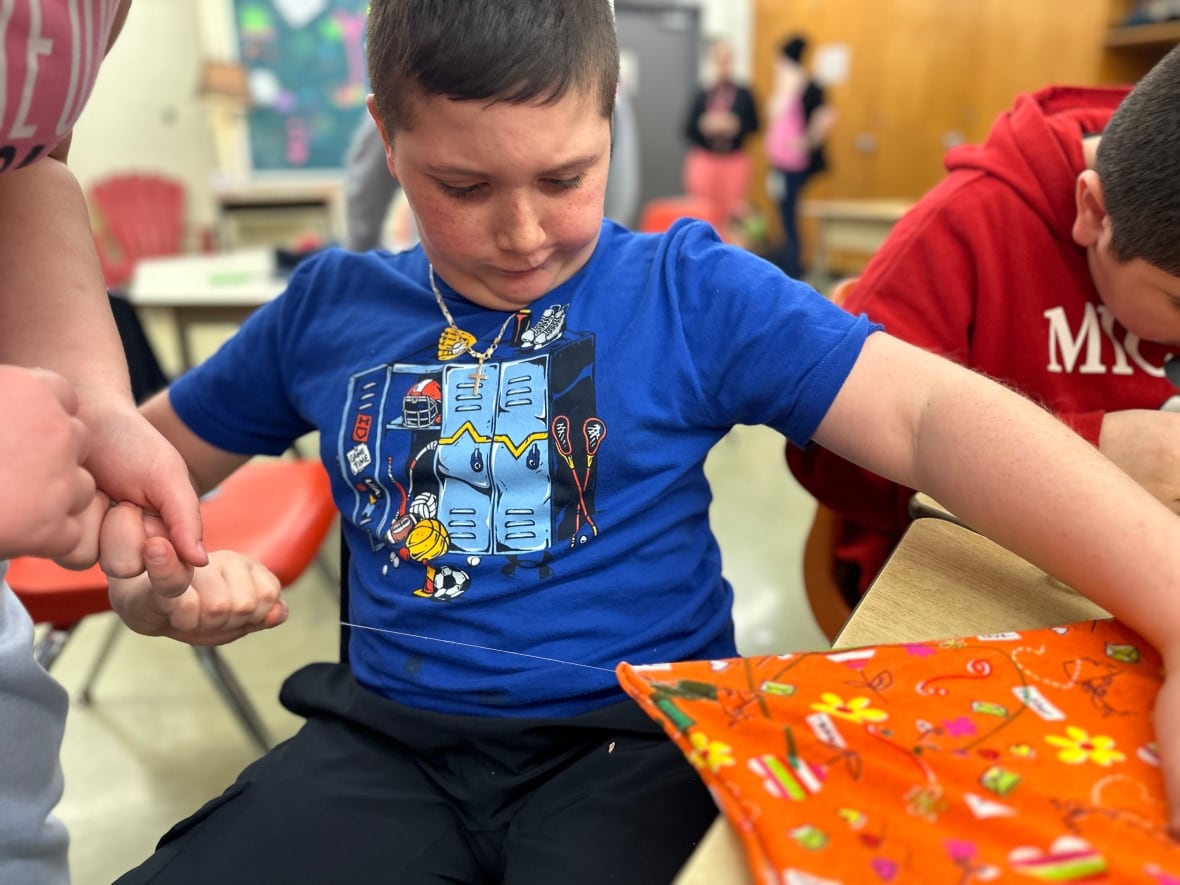 A little boy sews, pulling a needle and thread through an orange piece of cloth. 