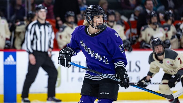 A hockey player in a purple jersey looks at something, with another team's player and a ref in the background.