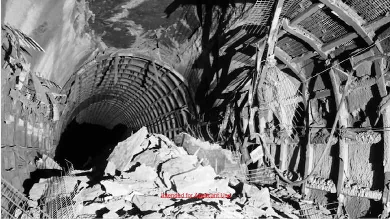 A black-and-white image of a pile of rocks in the middle of a dark tunnel, lit up by the flash of a camera, with bent steel hanging from the roof.