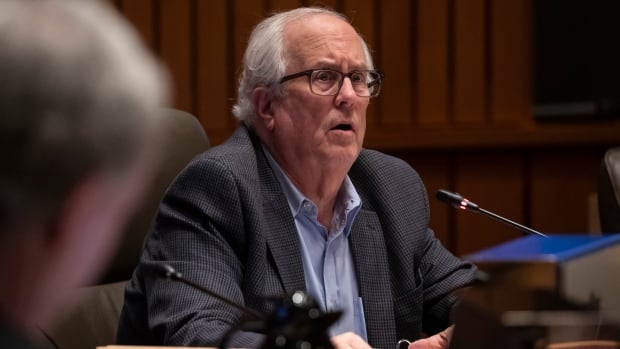 An senior man with white hair looks concerned as he sits in a city hall chamber.