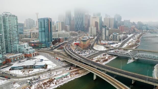 Shots from drone of Calgary Bow river and downtown core in December 2020. See city skyline and bridge. a city skyline is shown on a foggy day by a large river. a few roads pass overtop of the river.