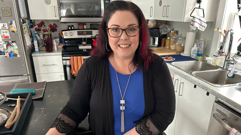 A woman with long dark hair and a blue shirt stands in her kitchen.