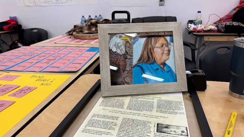 An Indigenous woman wearing glasses appears in a framed photo on a table. Behind it are scoreboards for a handgames tournament.