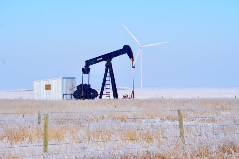 A wind turbine is pictured in the distance behind an oil pumpjack in a field.