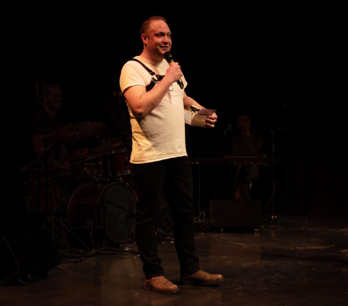 A man stands on stage holding a microphone, talking. The background is dark. He is wearing dark pants and a white shirt. 