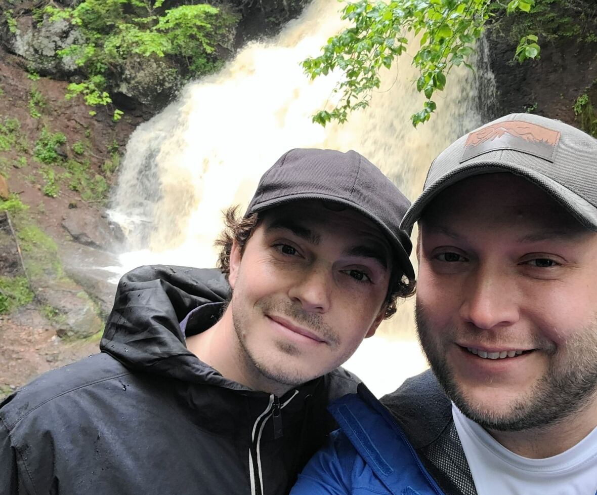 Two men pose happily outside in front of a waterfall. Both wearing rain jackets and baseball caps. 