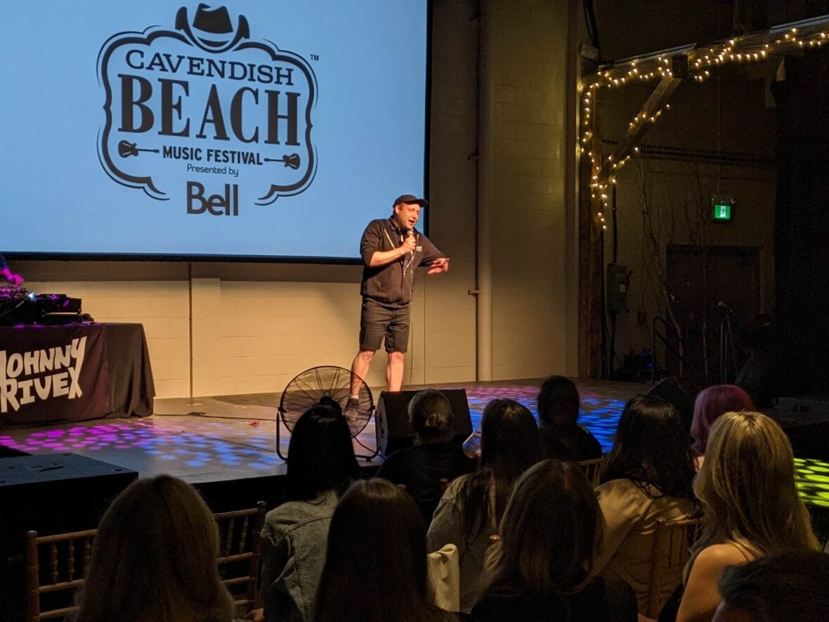 A man stand on a stage and talks into a microphone. He stands in front of a sign that says Cavendish Beach Festival. He is wearing black shorts, sweater and ball cap. 