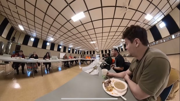 Arieh Smith can speak around 50 languages and has over six million followers on YouTube. He came to Northern Saskatchewan to learn the Cree language, promote it, and experience the culture of the Cree people. A man wearing a green shirt is sitting at a table with elders sitting around him