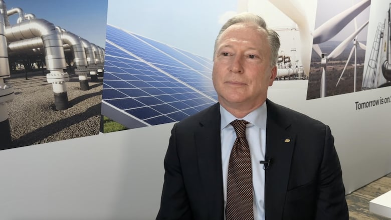 A man sits in front of a large poster showing natural gas pipes, solar panels and wind turbines.