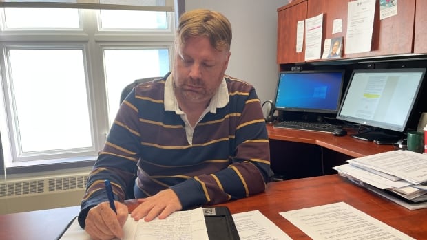A man wearing a brown and blue shirt writing at a desk filled with papers next to a computer