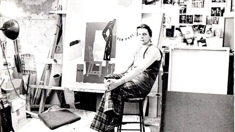 A person poses sitting on a stool in an art studio in front of a large easel, surrounded by art. 