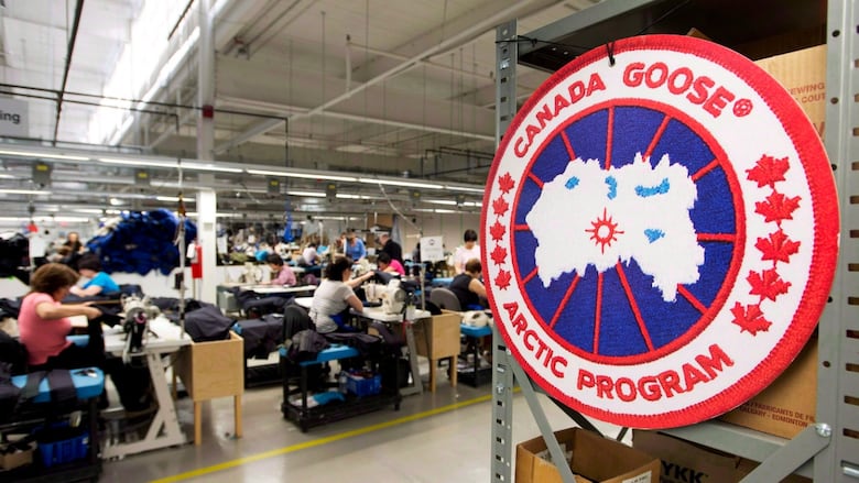 The Canada Goose logo is displayed in a factory setting with workers in the background.