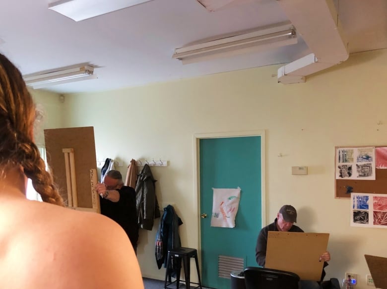A woman's hair and bare shoulder are visible while two people sit behind easels in the background.