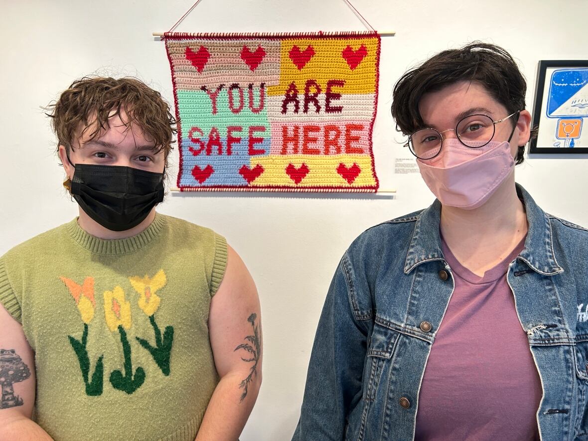 P.E.I. Transgender Network vice-chair Kal Ross, left, and executive director Lucky Fusca with a crocheted artwork behind them reading "you are safe here."