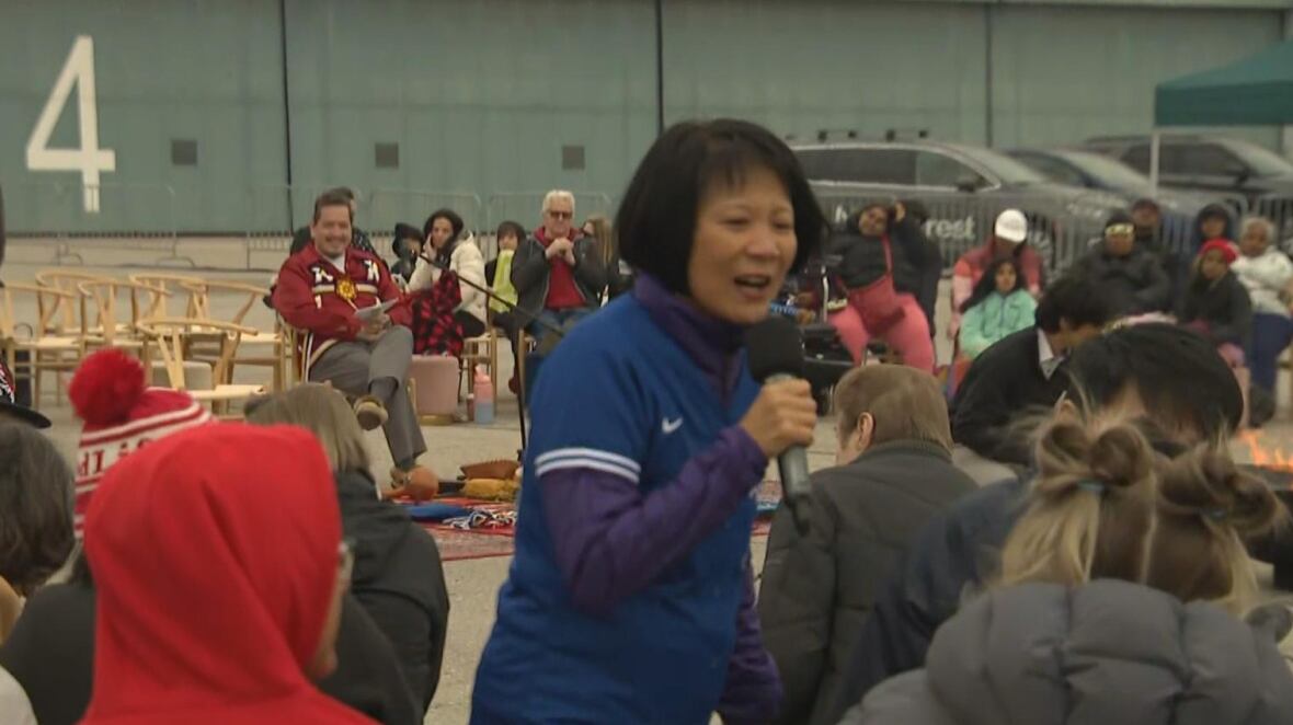 Woman wearing Blue Jays jersey speaks into a microphone in front of a crowd outside. 