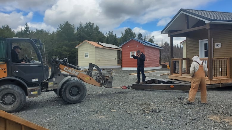 Name man wearing a black sweater and jeans stands on a trailer hitch loaded with a tiny home. He motions to a man driving a yellow construction vehicle to 'halt.'