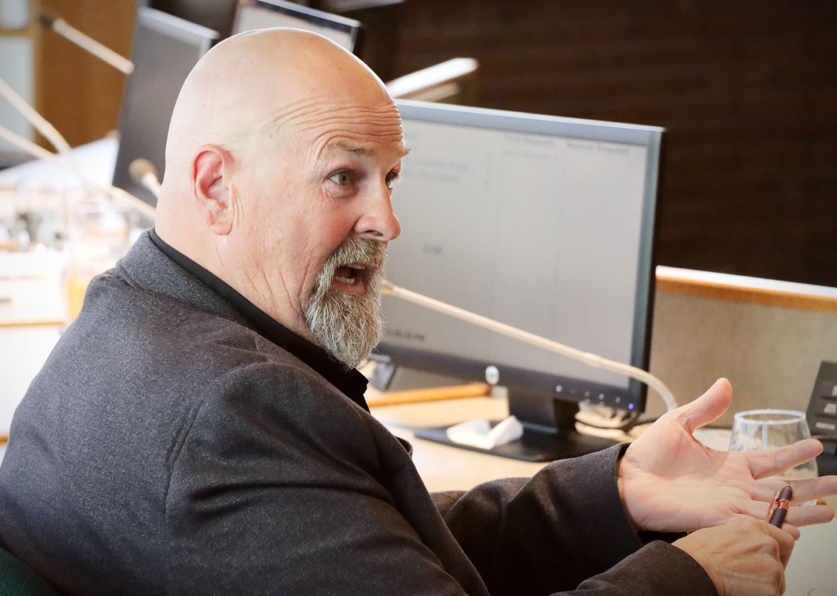 A bald man with a grey goatee and moustache wearing a dark suit jacket sits in front of a computer screen and gestures with his hands.