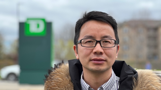 Customer who filed complaint against TD Bank refuses to sign gag order to get compensation; An East Asian man wearing glasses and a coat with fur trim stands in a parking lot in front of a blurred TD Bank logo\.
