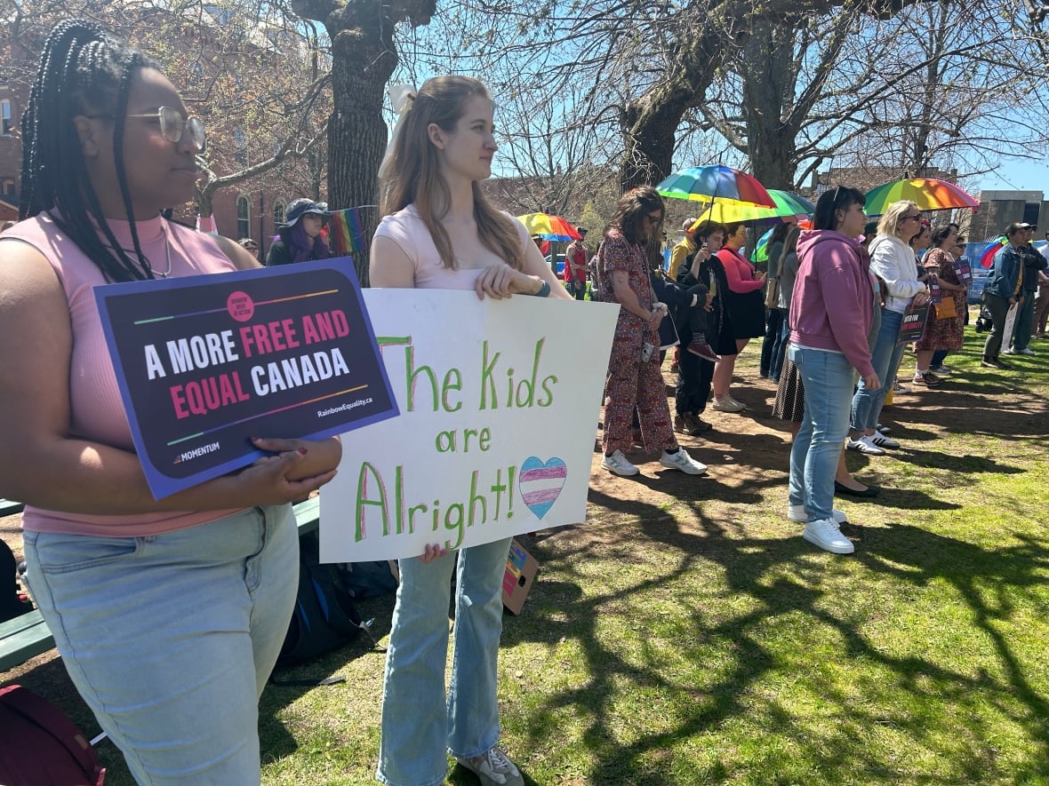 Two people hold signs in front of a group of more people holding signs and rainbow umbrellas.