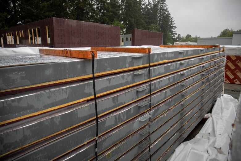 A stack of grey building panels is seen in the foreground, with a partially completed burgundy school building next to a forested area in the background.