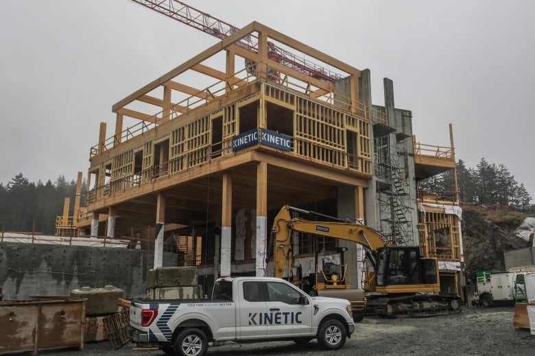 A white pick-up truck is seen in the foreground parked next to heavy construction equipment next to a partially built wood and concrete school structure.