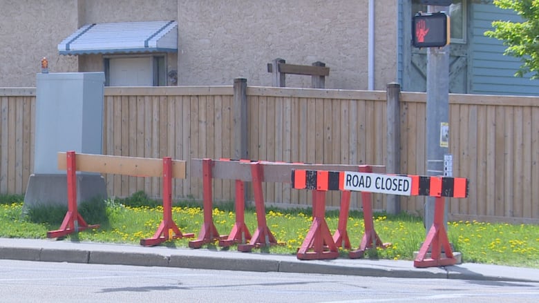 Orange-and-black 'Road closed' signs stand in the grass beside a sidewalk at four-way intersection. Above the signs is a streetlight showing the hand signal.