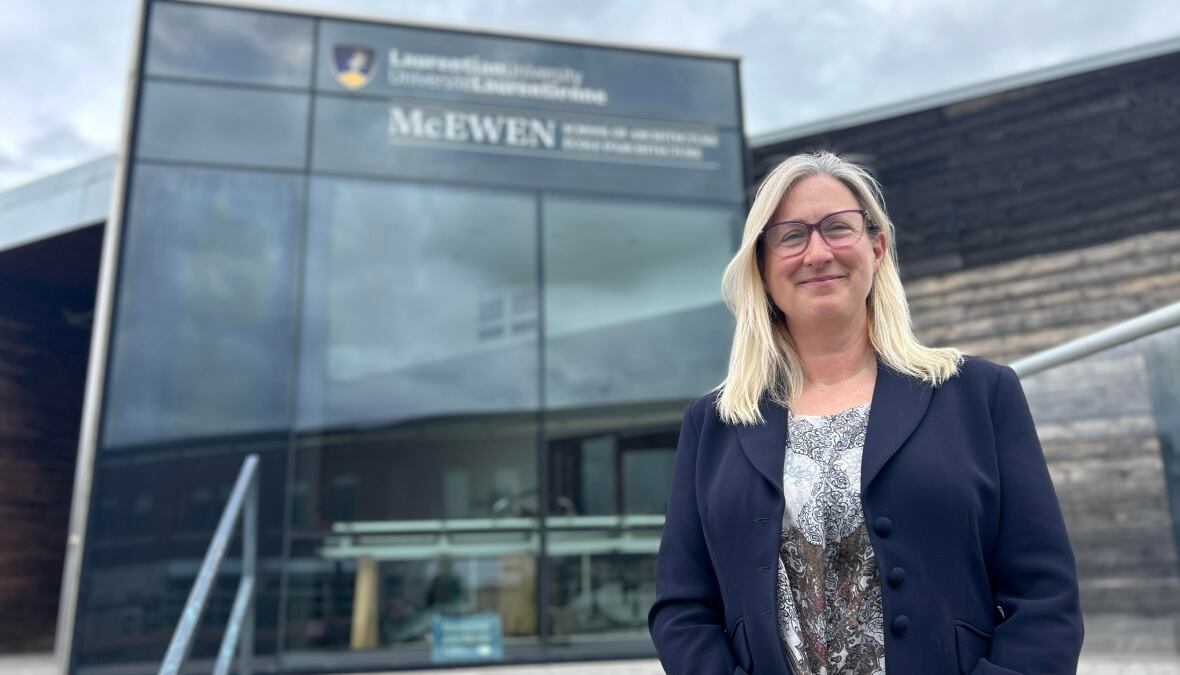 A smiling woman with blonde hair stands in front of the Laurentian University architecture school 