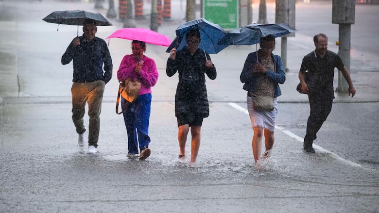 Five people cross an intersection in the rain. Four of them have umbrellas. At their feet water is pooling on the concrete and splashing as they walk.