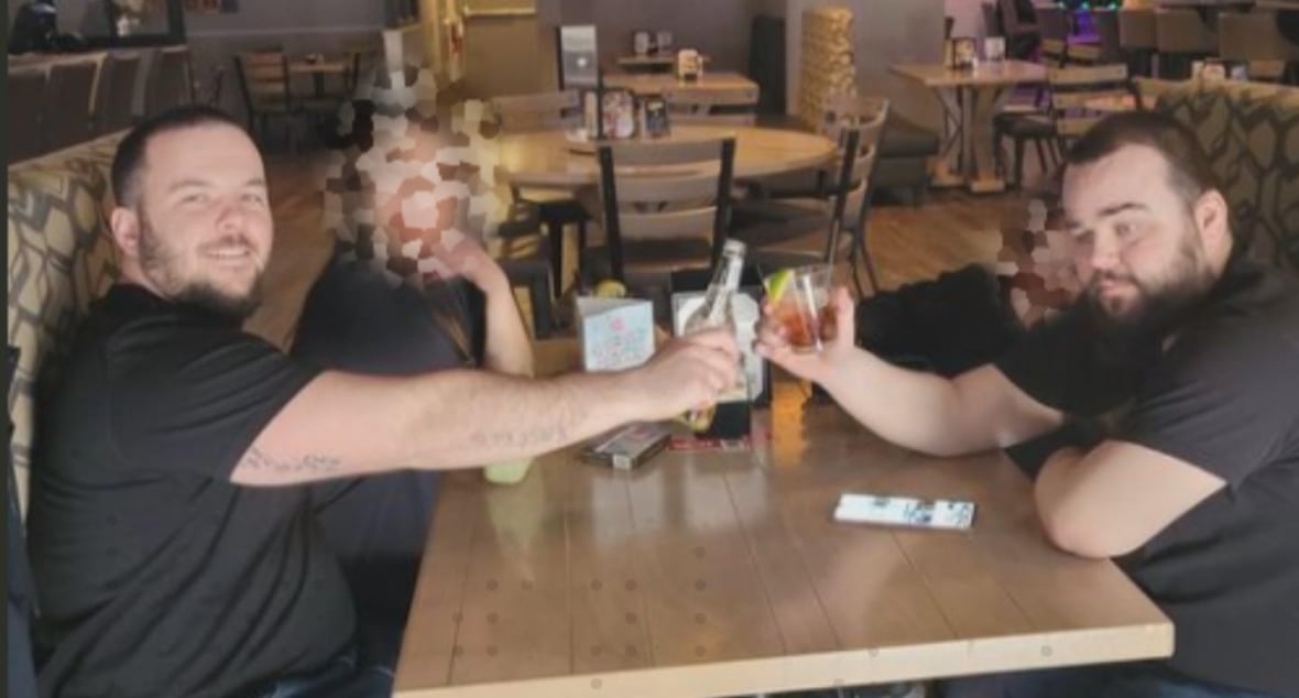 Two men in black t-shirts toast with drinks at a table at a restaurant.