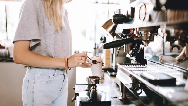 A stock image shows an employee at a coffee shop. A youthful blond lady, wearing casual clothes stands next to a coffee machine in a cozy coffee shop.