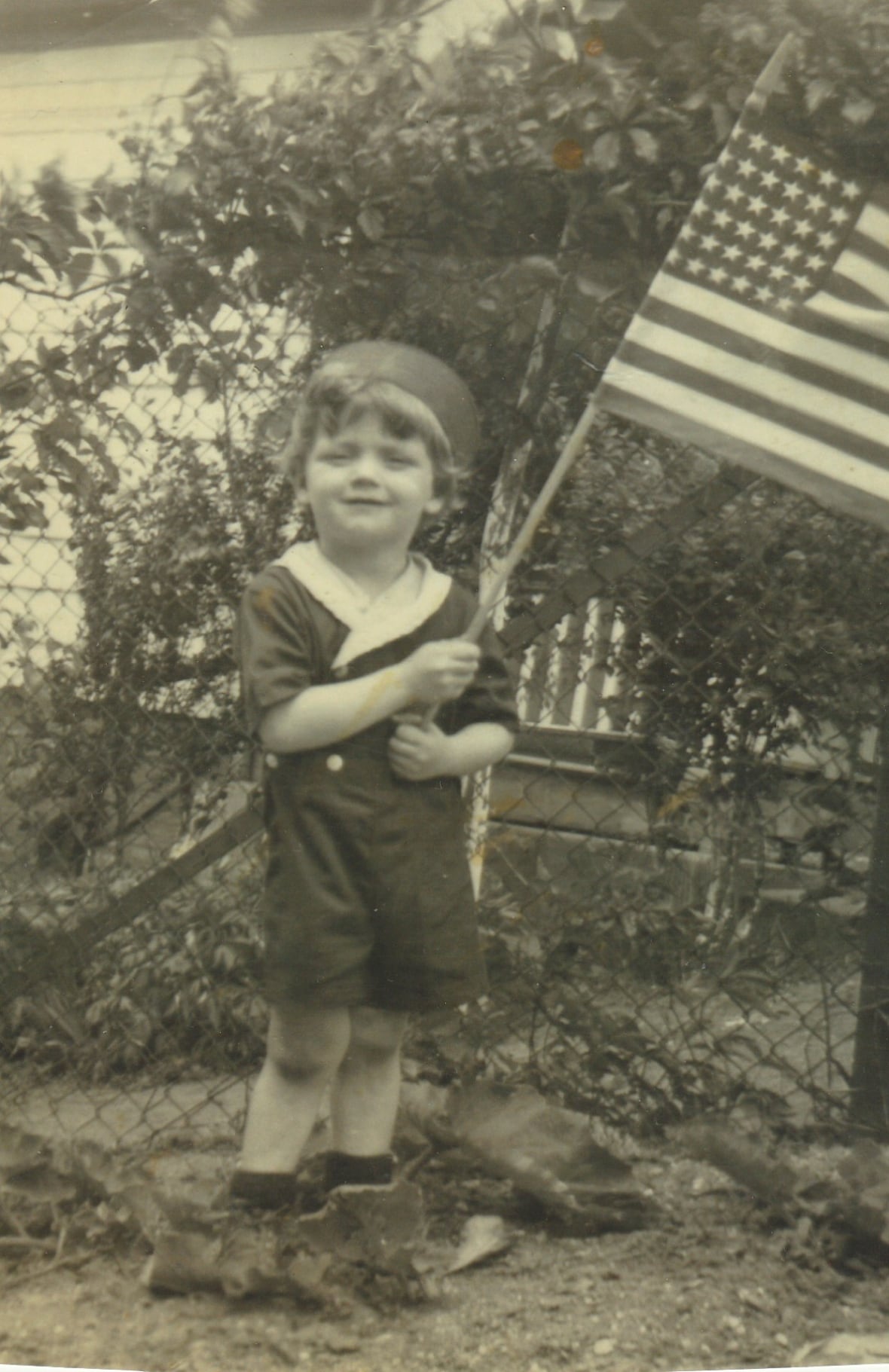 Black and white image of a little boy in a sailor's uniform, smiling and waving an American flag