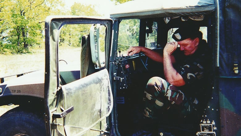A man sits at the wheel of Jeep with the door open, wearing military fatigues, and resting his head on his head.
