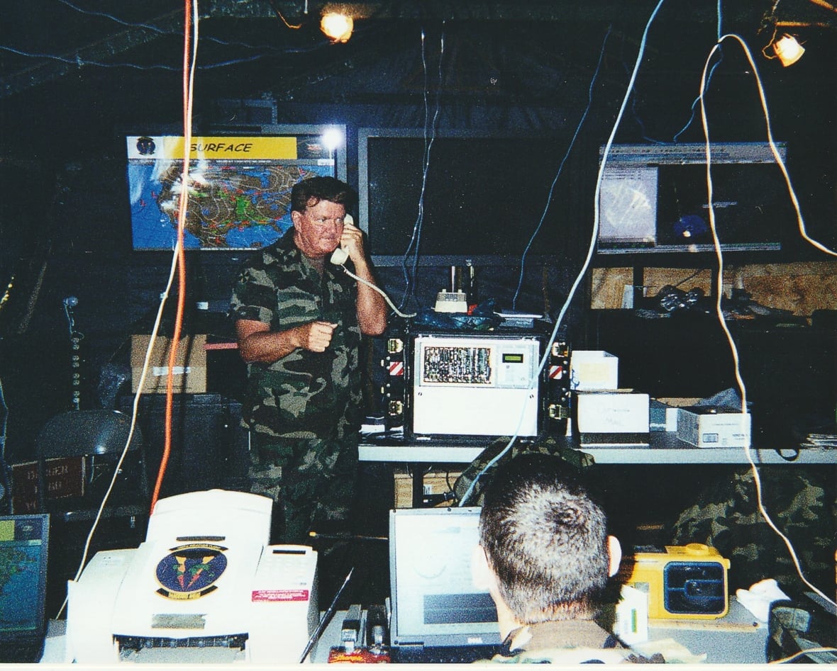 A man in military fatigues talks on a landline phone in a room full of computers, T screens and wires hanging from the ceilings.