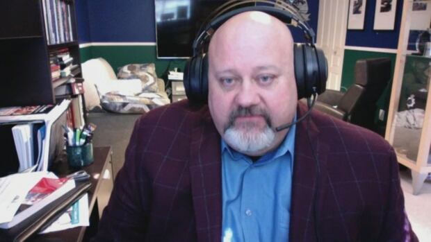 A bald white man wears headphones and speaks while looking at a computer in an office space.