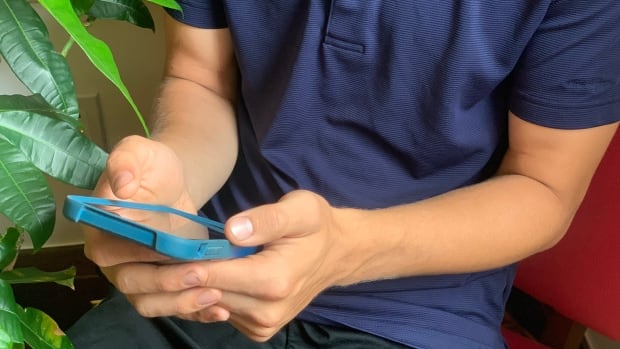 A young man's torso and hands are seen as he scrolls his phone while sitting on a bright pink chair next to a houseplant.