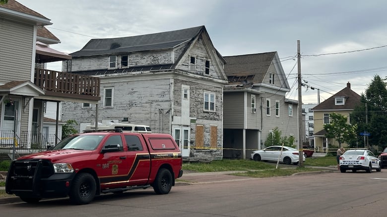 A red vehicle and police car are parked outside two large houses that are damaged from fire. Yellow tape stretches across the front of the buildings.