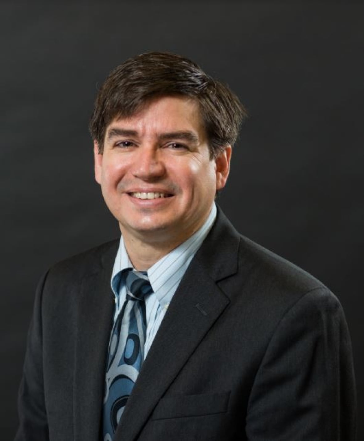 A white man with dark hair in a suit smiles at the camera